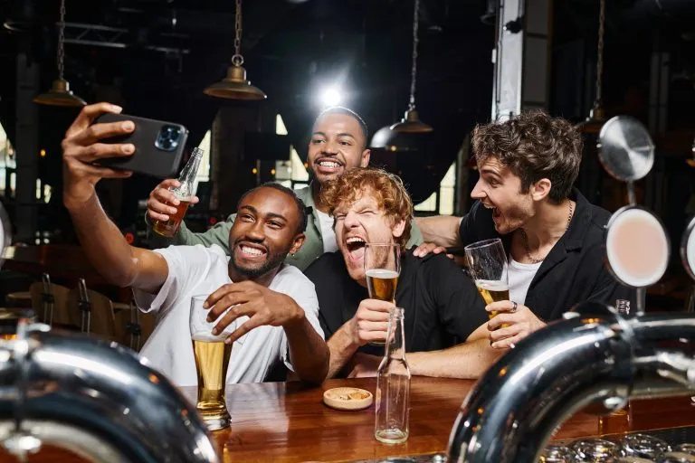 Des hommes multiculturels heureux prennent un selfie avec leur smartphone tout en buvant de la bière dans un bar, lors d'un enterrement de vie de garçon.